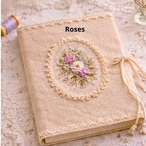 May include: A beige fabric journal with the word "Roses" printed on the cover. The journal features an embroidered floral design in a circular frame, with pink and purple flowers. The edges are trimmed with lace, and it has a ribbon tie closure.
