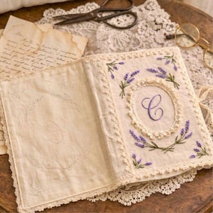 May include: An open, off-white fabric journal with a lavender floral embroidery design and the letter "C" in the center. The journal is trimmed with lace and has a matching embroidered design on the inside cover. Vintage-style.