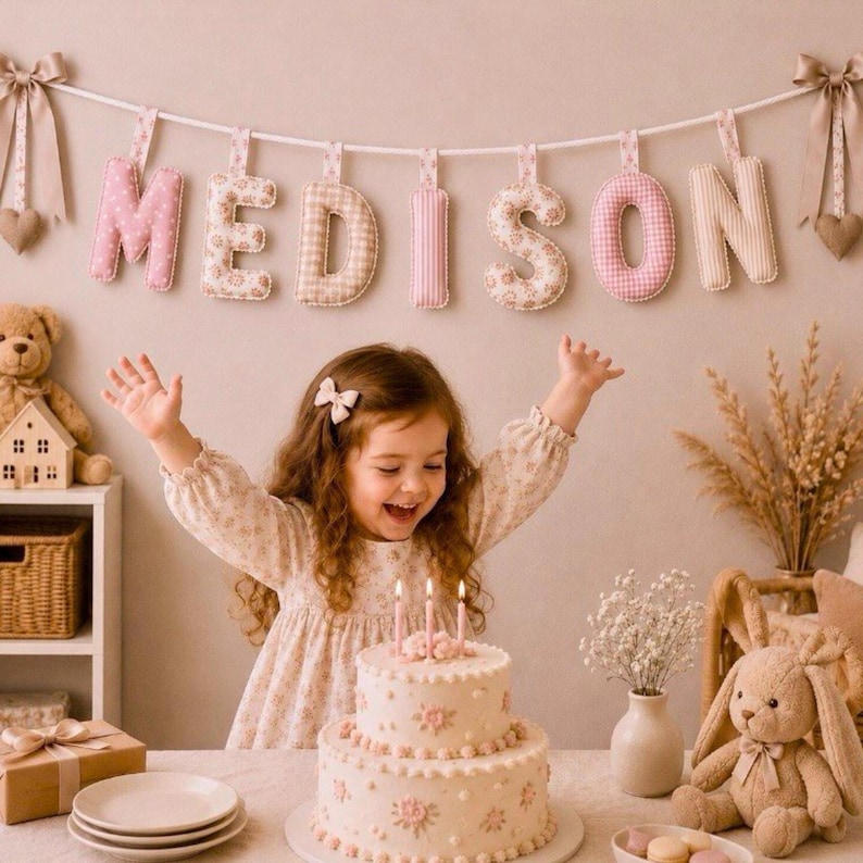 May include: A two-tiered birthday cake with lit candles sits in front of a young girl with her arms raised. A banner spelling "MEDISON" hangs above. The letters are in various floral and patterned fabrics. A teddy bear and a stuffed bunny are also visible.