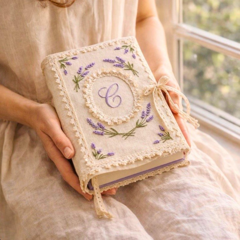 May include: A beige book with a lavender floral design. The cover features embroidered lavender sprigs, a central monogram, and a lace trim. A ribbon is tied on the side. The book is held in someone's hands.