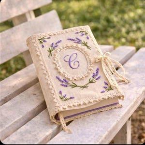 May include: A beige journal with embroidered lavender sprigs and a central monogram "C" in purple. The cover is trimmed with lace and tied with a cream-colored ribbon. The journal is resting on a weathered wooden bench.