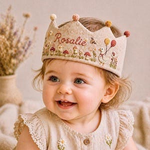 Peut inclure: Une couronne en feutre beige brodée du nom "Rosalie", de fleurs et de champignons. La couronne a un bord festonné et est ornée de petits pompons colorés. Le bébé porte une robe beige avec des broderies de marguerites.
