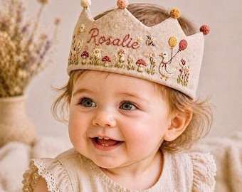 May include: A beige felt crown embroidered with the name "Rosalie", flowers, and mushrooms. The crown has a scalloped edge and is adorned with small, colorful pom-poms. The baby is wearing a beige dress with daisy embroidery.