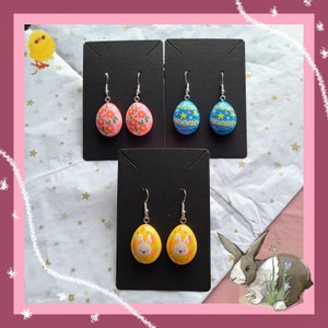 Puede incluir: Tres pares de pendientes de huevo de Pascua hechos a mano. Un par es rosa con diseños florales, un par es azul con diseños geométricos y un par es amarillo con una cara de conejo.