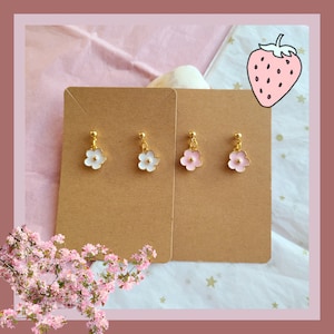 Pode incluir: Um par de brincos de flores douradas com esmalte branco e rosa. Os brincos são exibidos em um cartão marrom com fundo branco.