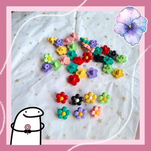 Pode incluir: Uma coleção de botões coloridos em forma de flor em várias cores, incluindo vermelho, amarelo, verde, roxo e preto. Os botões estão espalhados em uma superfície branca com padrões de estrelas douradas. Um personagem de desenho animado segurando uma flor está no canto inferior esquerdo.