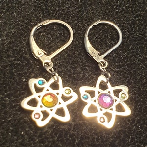 Może przedstawiać: Para srebrnych kolczyków z kolorowym wzorem atomu. Każdy kolczyk przedstawia srebrny atom z centralnym kryształem i mniejszymi kryształami wokół krawędzi.