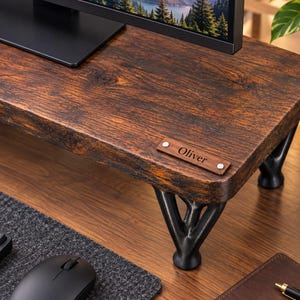 May include: A rectangular monitor stand with a dark brown wood grain finish and four black metal legs. A small rectangular wooden plaque is attached to the front right corner, featuring the engraved name Oliver in a dark serif font.