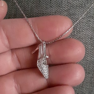 Pode incluir: Um colar de prata com um pingente de sapato de cristal transparente. O sapato é adornado com pequenos cristais transparentes.