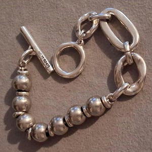 May include: A silver-toned bracelet featuring a toggle clasp with the text UNOde50 engraved on the bar. The design consists of a series of smooth, spherical beads on one side and a sequence of irregular, elongated oval links on the other side.