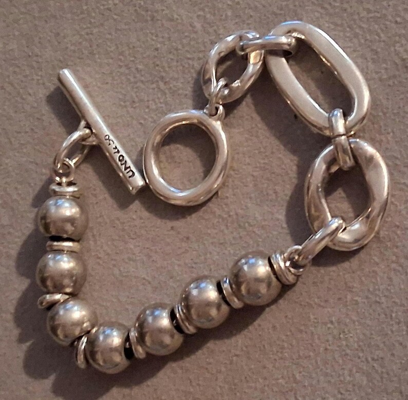 May include: A silver-toned bracelet featuring a toggle clasp with the text UNOde50 engraved on the bar. The design consists of a series of smooth, spherical beads on one side and a sequence of irregular, elongated oval links on the other side.
