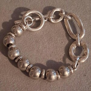 May include: A silver-toned bracelet featuring a series of smooth, spherical beads alternating with small flat spacers. The bracelet includes a toggle clasp with a circular ring and a T-bar, connected to two larger, irregular oval-shaped links.