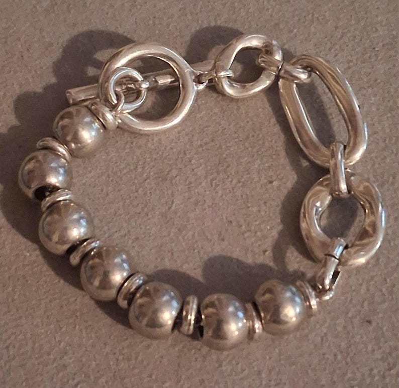May include: A silver-toned bracelet featuring a series of smooth, spherical beads alternating with small flat spacers. The bracelet includes a toggle clasp with a circular ring and a T-bar, connected to two larger, irregular oval-shaped links.