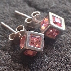 May include: A pair of stud earrings featuring a silver-toned cube frame. Each cube contains a faceted, square-cut pink gemstone on its visible faces. The earrings include standard silver-toned posts and butterfly-style backings for secure attachment.