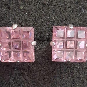 May include: A pair of square-shaped stud earrings. Each earring features a pink, faceted, square-cut gemstone set in a silver metal setting. The gemstones are divided into a grid pattern, creating a textured appearance.