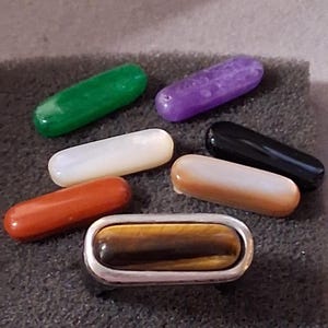 May include: A selection of polished gemstone beads in shades of green, purple, white, orange, black, and a tiger's eye stone set in silver-coloured metal. The oblong beads are displayed on a dark grey surface.