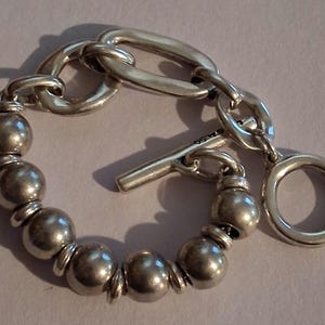 May include: A silver-toned bracelet featuring a series of smooth, spherical beads separated by small flat discs. The bracelet includes a toggle clasp mechanism with a T-bar and a circular ring, connected to a section of elongated oval chain links.