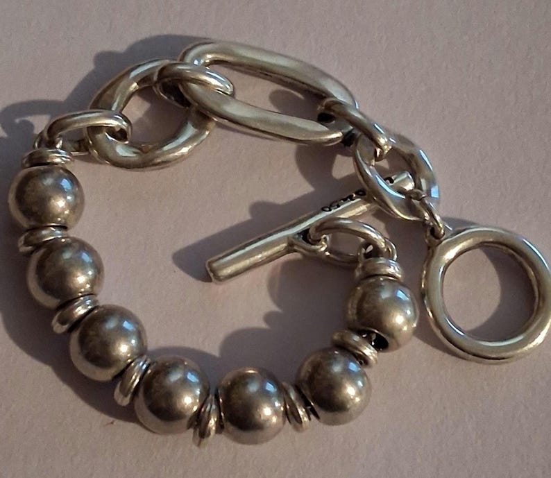 May include: A silver-toned bracelet featuring a series of smooth, spherical beads separated by small flat discs. The bracelet includes a toggle clasp mechanism with a T-bar and a circular ring, connected to a section of elongated oval chain links.