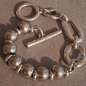 May include: A silver-toned bracelet featuring a series of smooth, spherical beads separated by small flat spacers. The bracelet includes a toggle clasp mechanism with a circular ring and a straight bar, connected to a section of larger oval links.