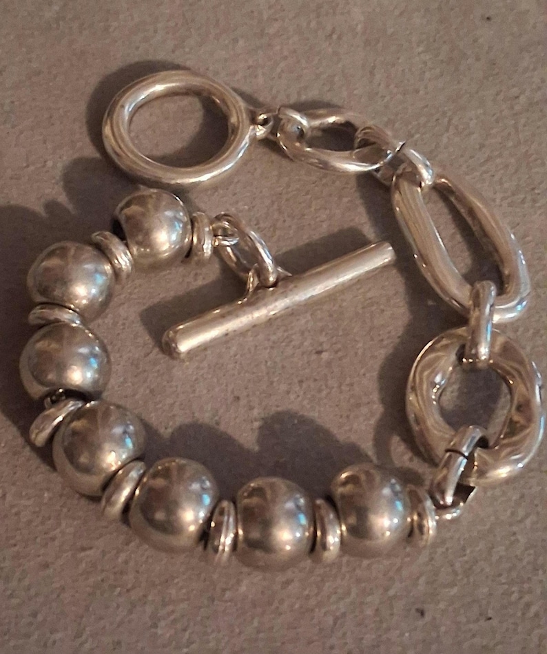 May include: A silver-toned bracelet featuring a series of smooth, spherical beads separated by small flat spacers. The bracelet includes a toggle clasp mechanism with a circular ring and a straight bar, connected to a section of larger oval links.