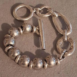 May include: A silver-toned bracelet featuring a series of smooth, spherical beads alternating with small flat rings, connected to a toggle clasp with a T-bar and a circular ring, alongside two larger, irregular oval-shaped links on one side.