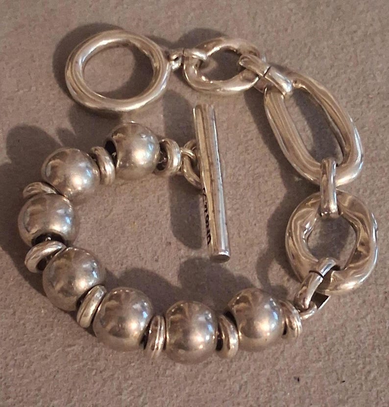May include: A silver-toned bracelet featuring a series of smooth, spherical beads alternating with small flat rings, connected to a toggle clasp with a T-bar and a circular ring, alongside two larger, irregular oval-shaped links on one side.