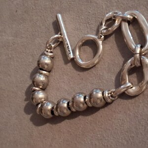 May include: A silver-toned bracelet featuring a series of smooth, spherical beads on one side and two large, elongated oval links on the other. The piece is secured with a toggle clasp consisting of a T-bar and a circular ring, with text visible on the T-bar.