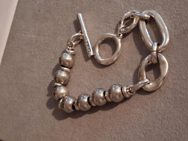 May include: A silver-toned bracelet featuring a series of smooth, spherical beads on one side and two large, elongated oval links on the other. The piece is secured with a toggle clasp consisting of a T-bar and a circular ring, with text visible on the T-bar.