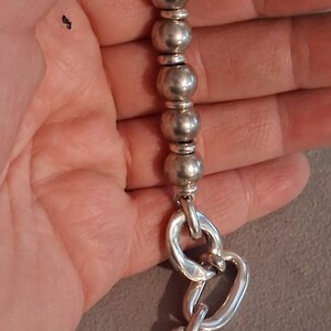 May include: A silver-toned bracelet featuring a series of smooth, spherical beads separated by smaller, flat disc spacers, transitioning into a section of large, irregular, oval-shaped chain links with a polished, reflective metallic finish.
