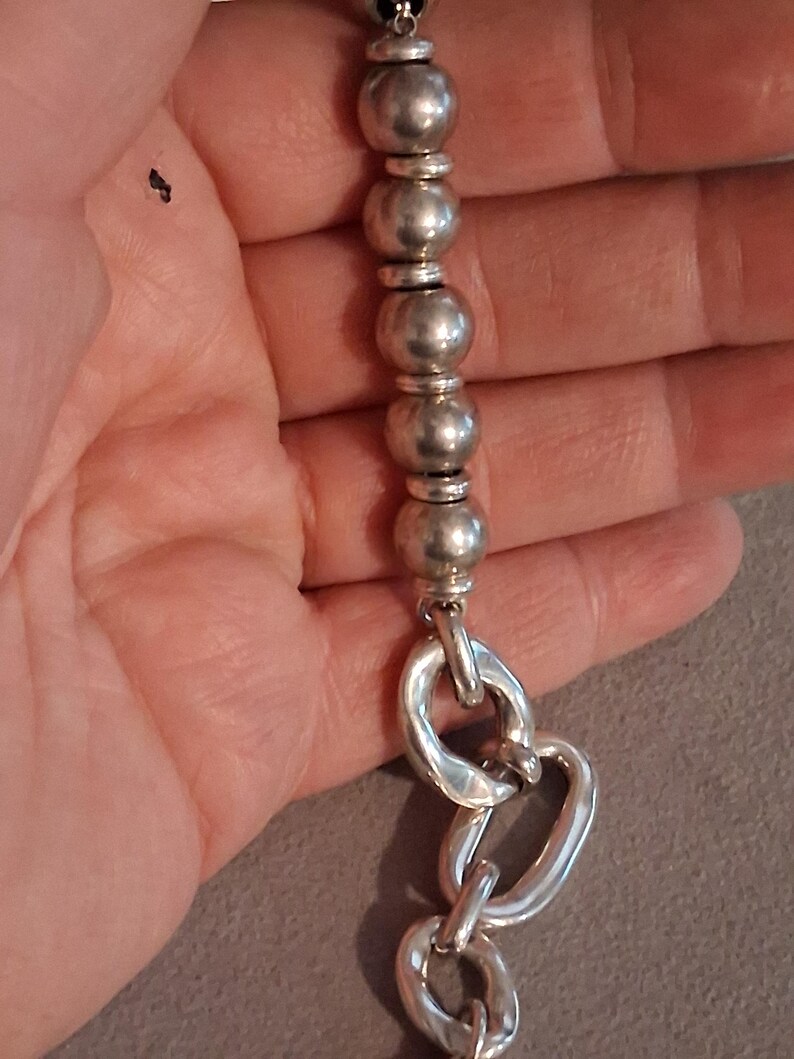 May include: A silver-toned bracelet featuring a series of smooth, spherical beads separated by smaller, flat disc spacers, transitioning into a section of large, irregular, oval-shaped chain links with a polished, reflective metallic finish.