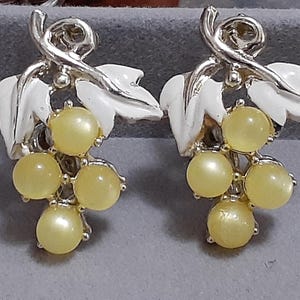 Puede incluir: Un par de pendientes de tono plateado con hojas de esmalte blanco y uvas de vidrio amarillo.