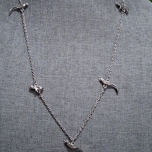 May include: A silver-toned necklace featuring a delicate chain design. The necklace is adorned with several small, bird-shaped pendants. The birds are evenly spaced along the chain, creating a graceful and elegant look. The necklace is displayed against a gray background.