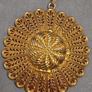 May include: A gold-colored pendant with a floral design. The pendant features a central spiral design surrounded by a series of petal-like shapes with intricate cut-out patterns. The pendant has a small loop at the top for a chain.