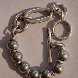 May include: A silver-toned bracelet featuring a series of round beads separated by small flat discs, connected to a large oval link and a circular ring. A T-bar toggle clasp is attached, which is engraved with the text UNOde50 in black lettering.