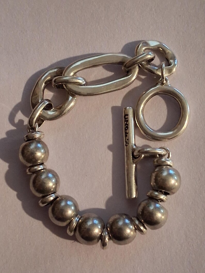 May include: A silver-toned bracelet featuring a series of round beads separated by small flat discs, connected to a large oval link and a circular ring. A T-bar toggle clasp is attached, which is engraved with the text UNOde50 in black lettering.