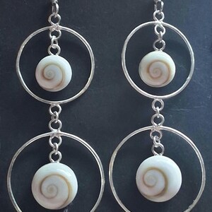 May include: A pair of silver-toned drop earrings featuring two stacked circular hoops. Each hoop contains a small, round, white shell bead with a natural brown spiral pattern centered within the frame, suspended by silver-toned metal links.