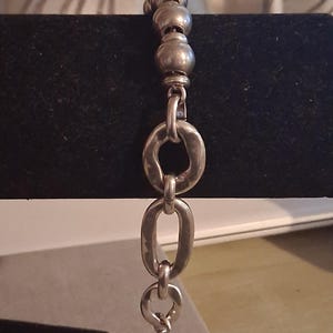May include: A silver-toned bracelet featuring a series of smooth, spherical beads at the top, transitioning into a sequence of irregular, hammered-texture oval links that connect to a circular clasp at the bottom end of the chain.