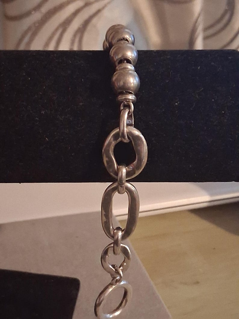 May include: A silver-toned bracelet featuring a series of smooth, spherical beads at the top, transitioning into a sequence of irregular, hammered-texture oval links that connect to a circular clasp at the bottom end of the chain.