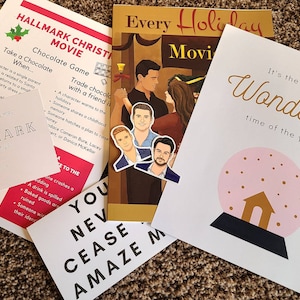 May include: A set of four cards with a Christmas theme. The cards feature a snow globe, a Hallmark Christmas movie game, a sticker of four men, and a card with the text "You Never Cease to Amaze Me".