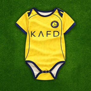 May include: Yellow baby bodysuit with navy blue trim and the letters KAFD printed on the front. The bodysuit has short sleeves and a round neckline. A circular emblem is on the upper chest.