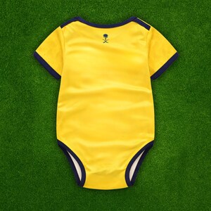 May include: A yellow baby onesie with navy blue trim around the neckline, sleeves, and leg openings. A small navy blue emblem is on the back. The onesie is made of a soft, satin-like material and is laid on a green surface.