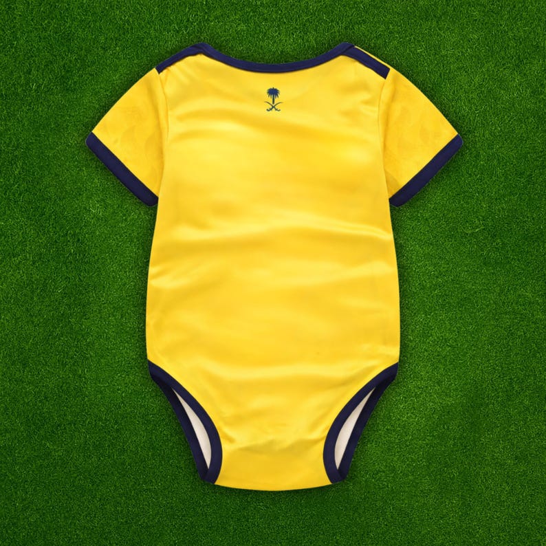 May include: A yellow baby onesie with navy blue trim around the neckline, sleeves, and leg openings. A small navy blue emblem is on the back. The onesie is made of a soft, satin-like material and is laid on a green surface.