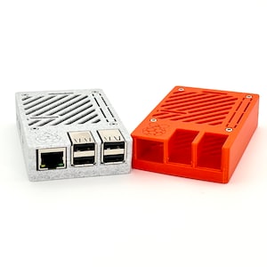Op de afbeelding: Twee Raspberry Pi-behuizingen, een grijs en een oranje, met een rasterpatroon aan de voorkant en een Raspberry Pi-logo op de oranje behuizing. Beide behuizingen hebben slots voor poorten en een ventilatieontwerp.