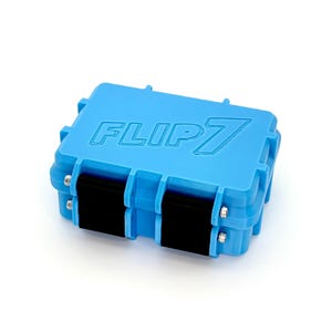 Op de afbeelding: Een lichtblauwe rechthoekige koffer met de tekst "FLIP 7" in reliëf op de bovenkant. De koffer heeft zwarte sluitingen en zilveren scharnieren. Op een witte achtergrond.