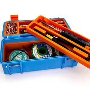 Könnte beinhalten: Eine blaue und orangefarbene Plastikwerkzeugbox mit einem Lötkolben, Draht und Lötmaterial im Inneren. Die Werkzeugbox hat einen Griff und einen Verschluss.