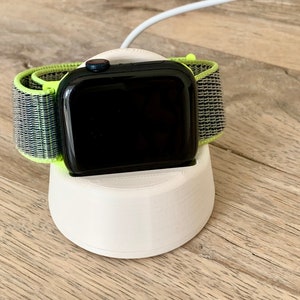 May include: A black smartwatch with a neon green and gray band rests on a white charging stand. The watch face is black, and a white charging cable is connected. The stand is on a wooden surface.