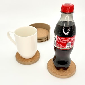 K&ouml;nnte beinhalten: Eine wei&szlig;e Keramiktasse und eine Coca-Cola-Flasche stehen auf runden Untersetzern aus Kork mit hellbraunem Rand. Zus&auml;tzliche Untersetzer sind in einem Halter gestapelt. Die Flasche hat einen roten Deckel.
