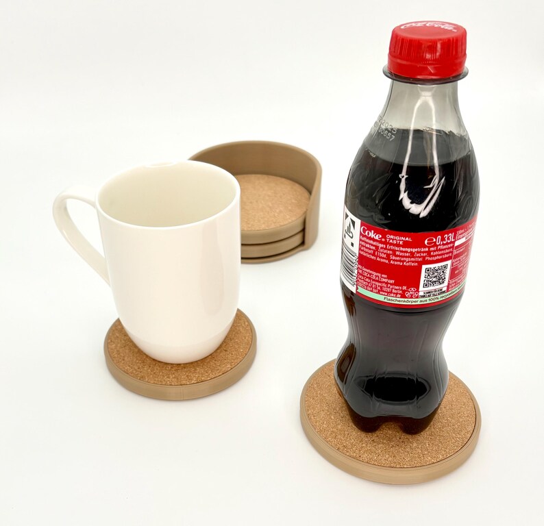 K&ouml;nnte beinhalten: Eine wei&szlig;e Keramiktasse und eine Coca-Cola-Flasche stehen auf runden Untersetzern aus Kork mit hellbraunem Rand. Zus&auml;tzliche Untersetzer sind in einem Halter gestapelt. Die Flasche hat einen roten Deckel.