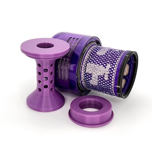 Puede incluir: Un filtro de aspiradora desmontado y sus accesorios. El filtro tiene forma cilíndrica con un exterior de malla. Los accesorios son de color morado claro, incluyendo un soporte con diseño de panal y un anillo circular.