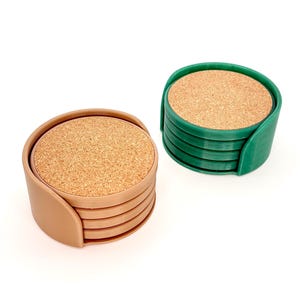 K&ouml;nnte beinhalten: Zwei Sets runde Untersetzer mit Korkeins&auml;tzen. Ein Set befindet sich in einem beigen Halter, das andere in einem gr&uuml;nen Halter. Jedes Set enth&auml;lt mehrere Untersetzer.